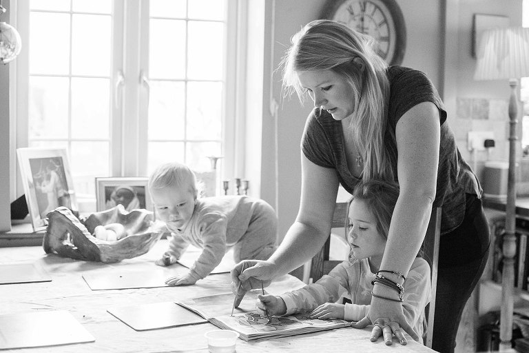 Mother helps young daughter with colouring as toddler climbs table.